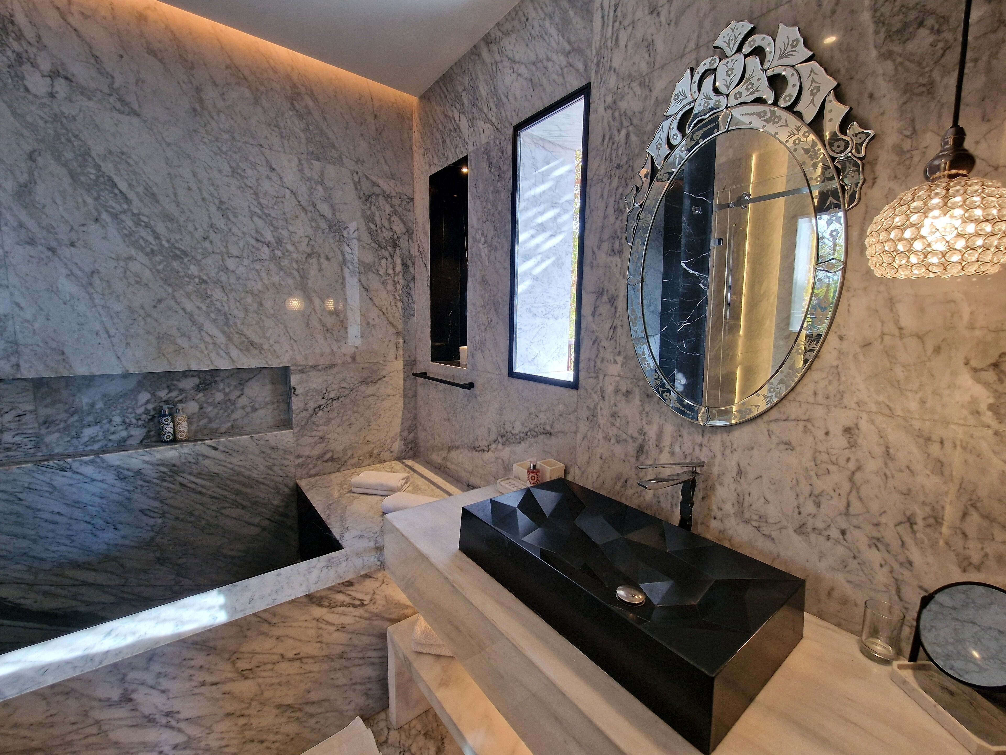 A modern bathroom with a marble wall, a freestanding bathtub, a black vanity with a mirror, and a hanging light fixture.