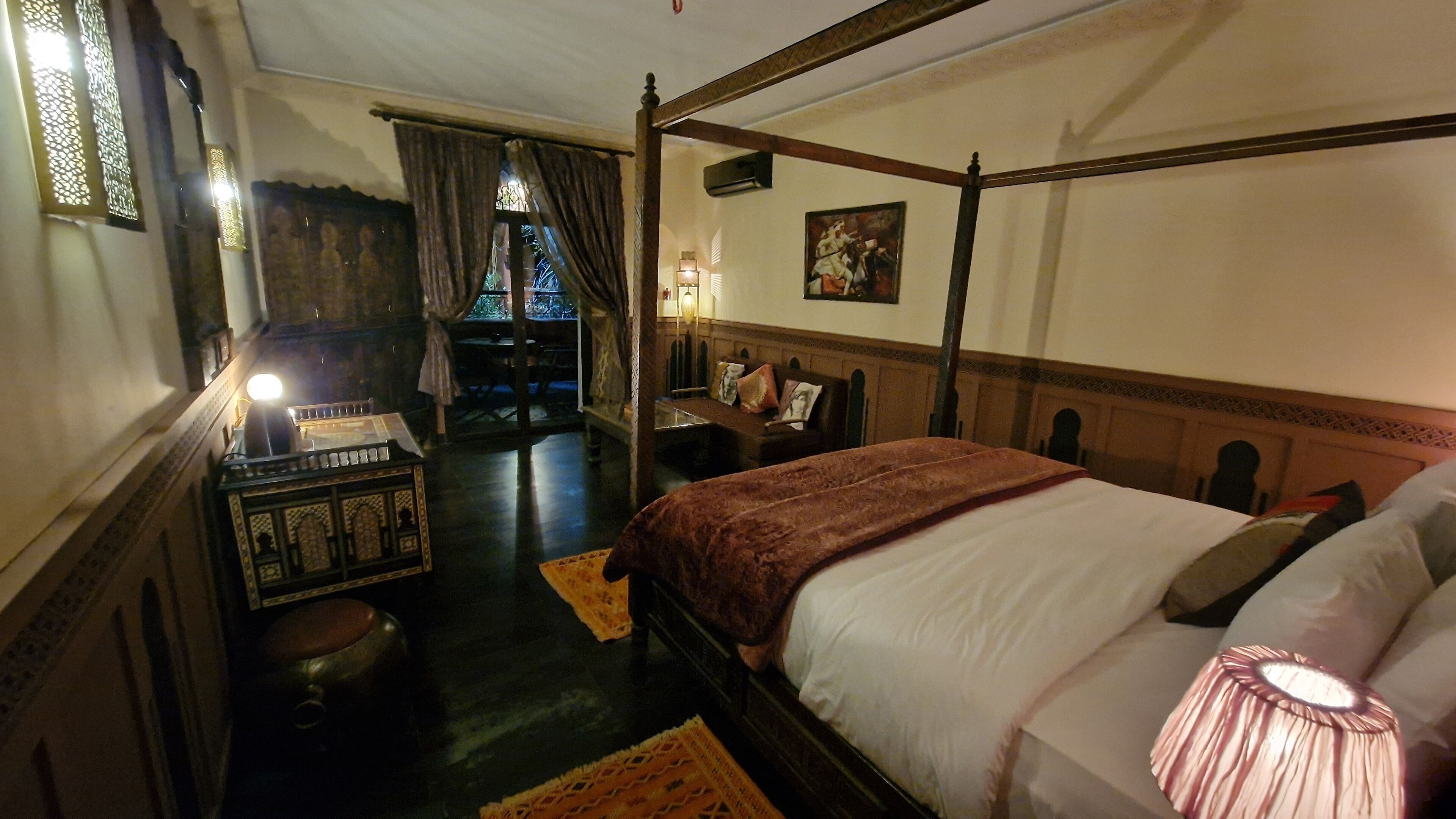 A traditional four-poster bed with a wooden headboard, a bedside table with a lamp, and a view of a courtyard through a window with curtains.