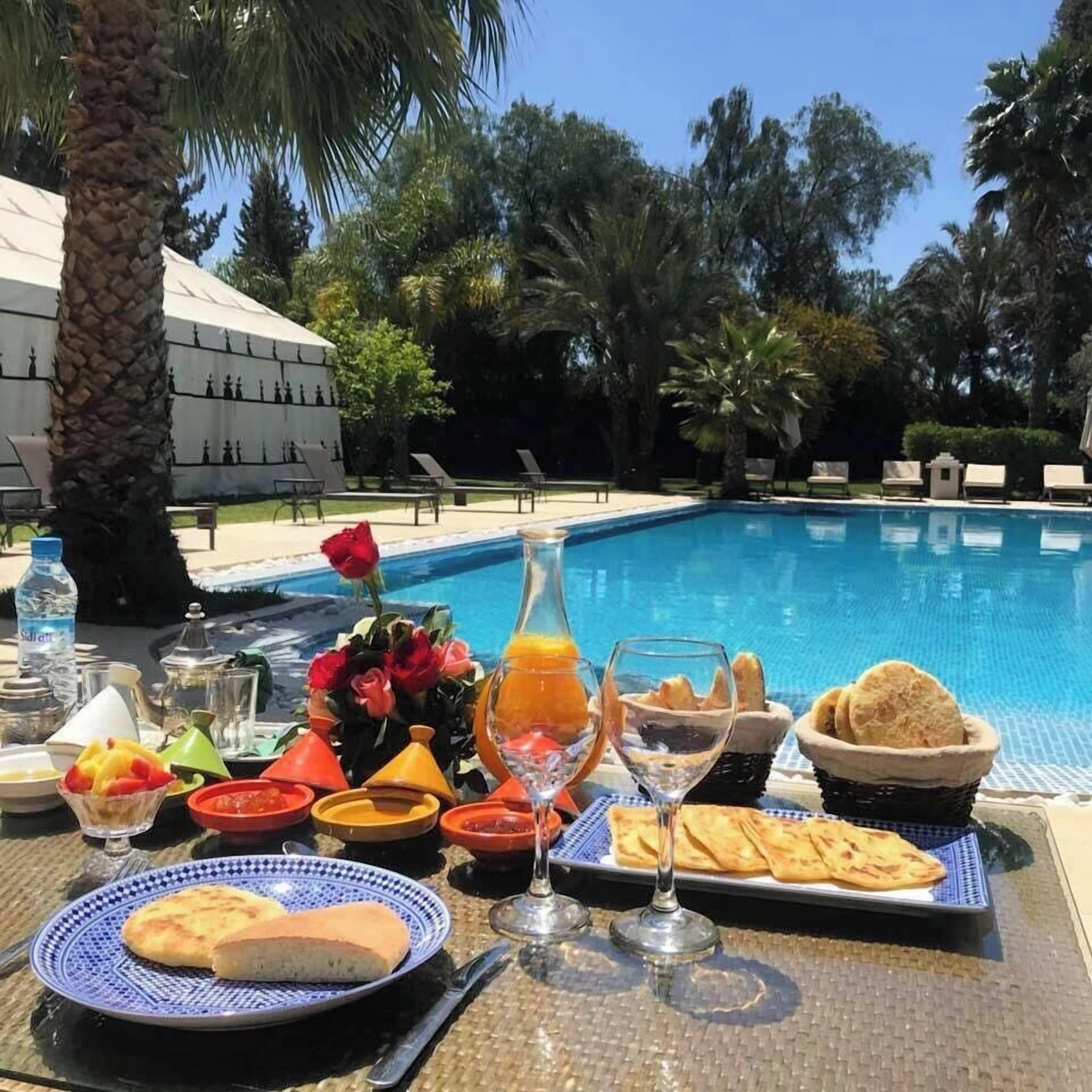 A breakfast setup by a pool with a table, plates, glasses, and a bottle of water.
