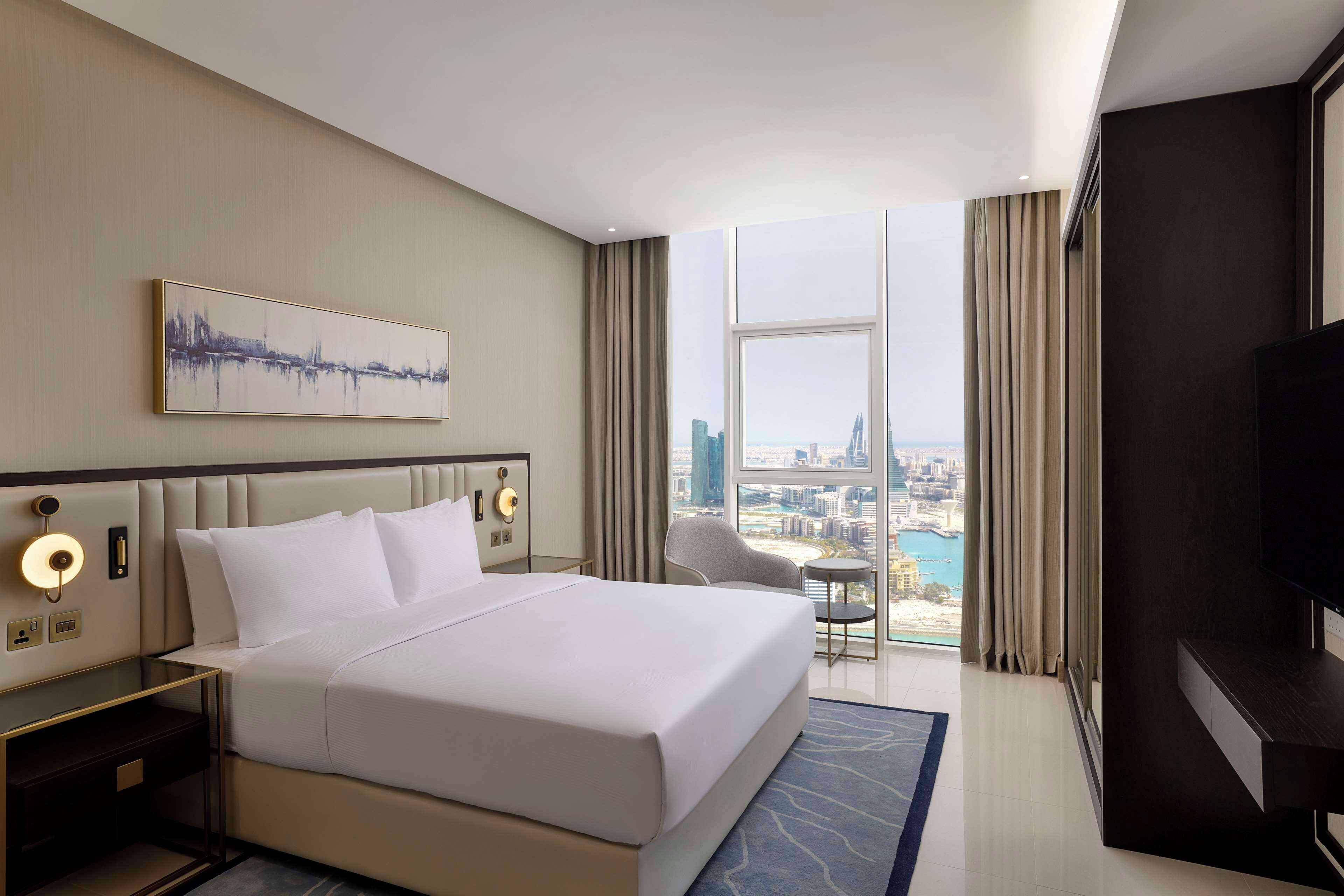 A hotel room with a large bed, a desk with a lamp, a chair, a TV, and a city view.