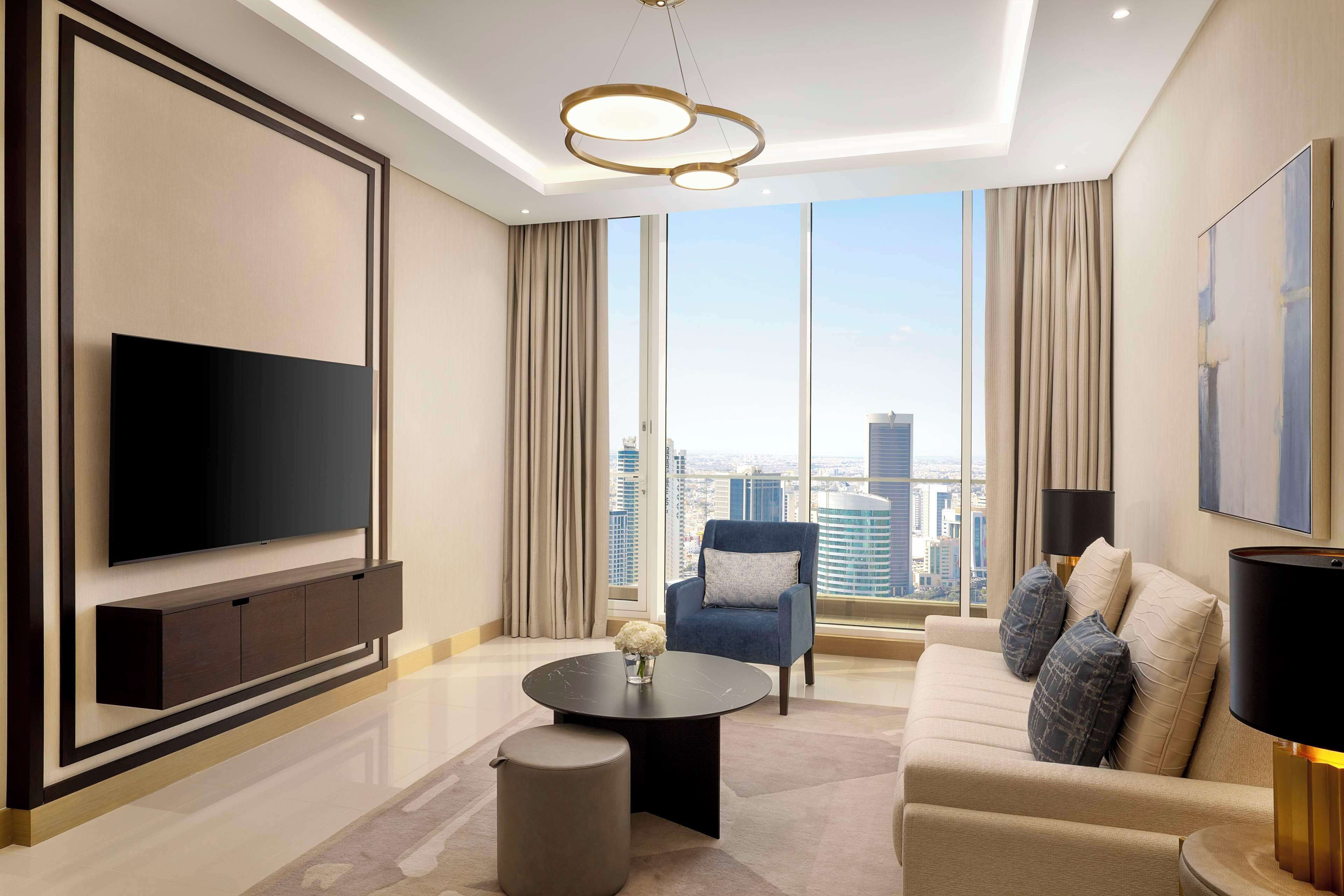 A modern living room with a flat-screen TV, a sofa, a chair, a coffee table, and a large window with a city view.