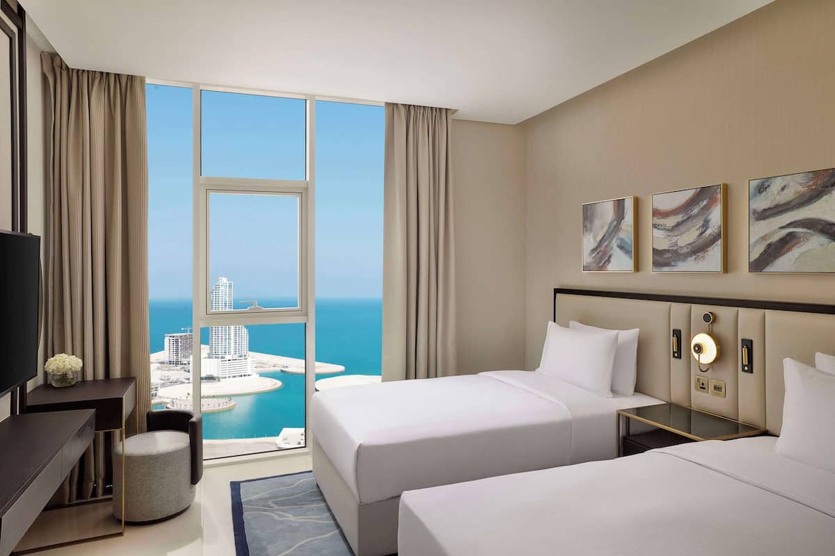 Habitación de hotel con dos camas, televisión, escritorio y vistas a la ciudad y al agua.