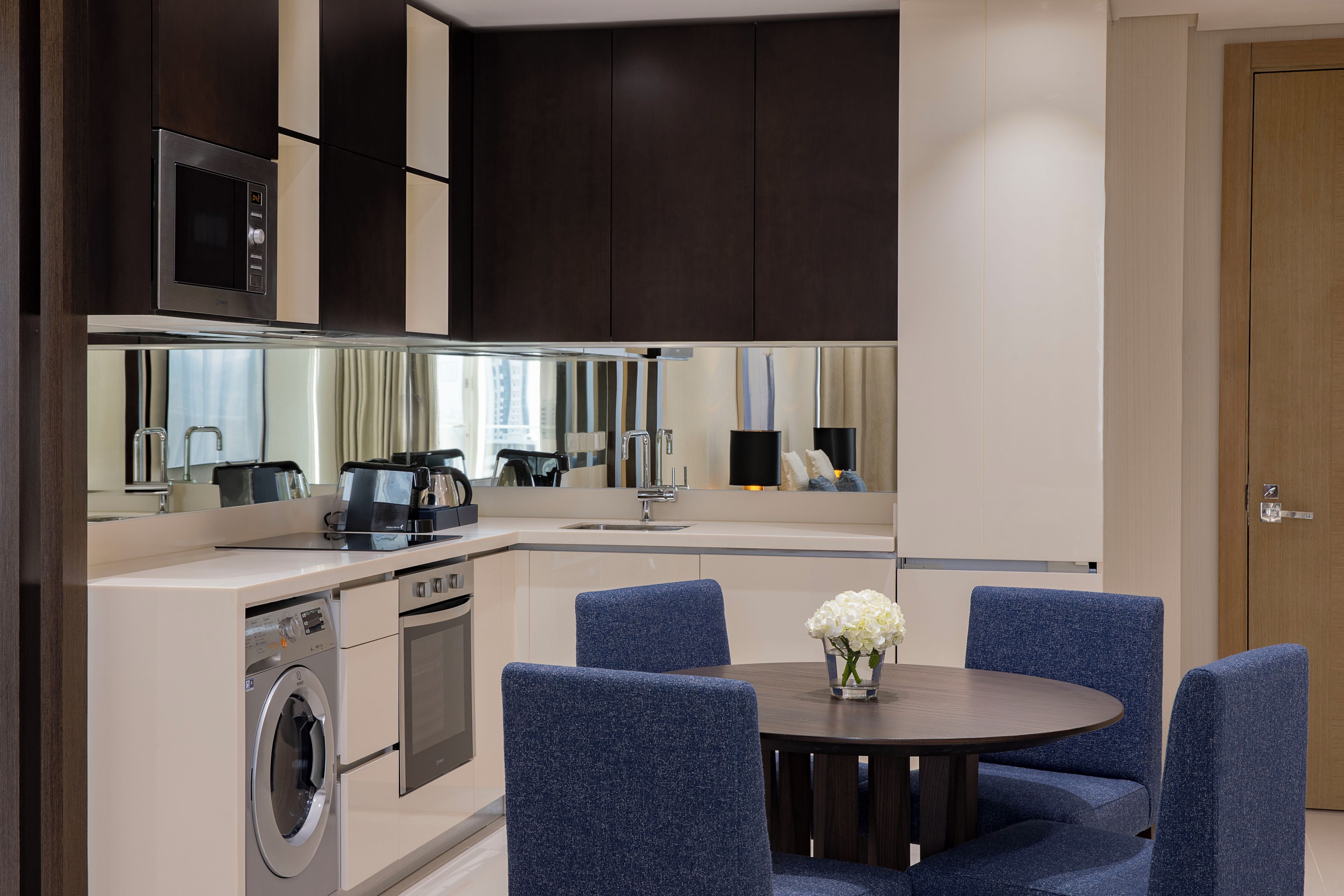 A modern kitchen with a built-in washer and dryer, a microwave, and a dining area with blue upholstered chairs and a round table.