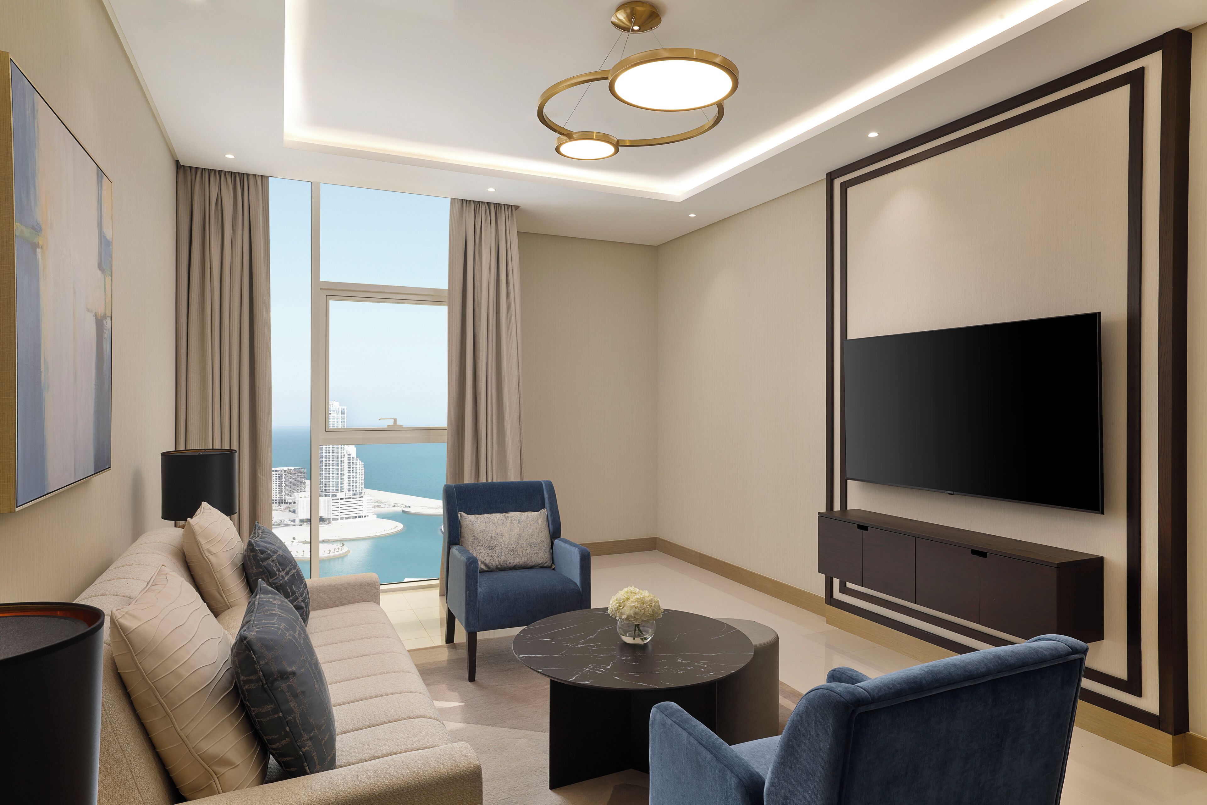 A modern living room with a sofa, armchairs, a coffee table, a TV, and a large window with a view of a cityscape.