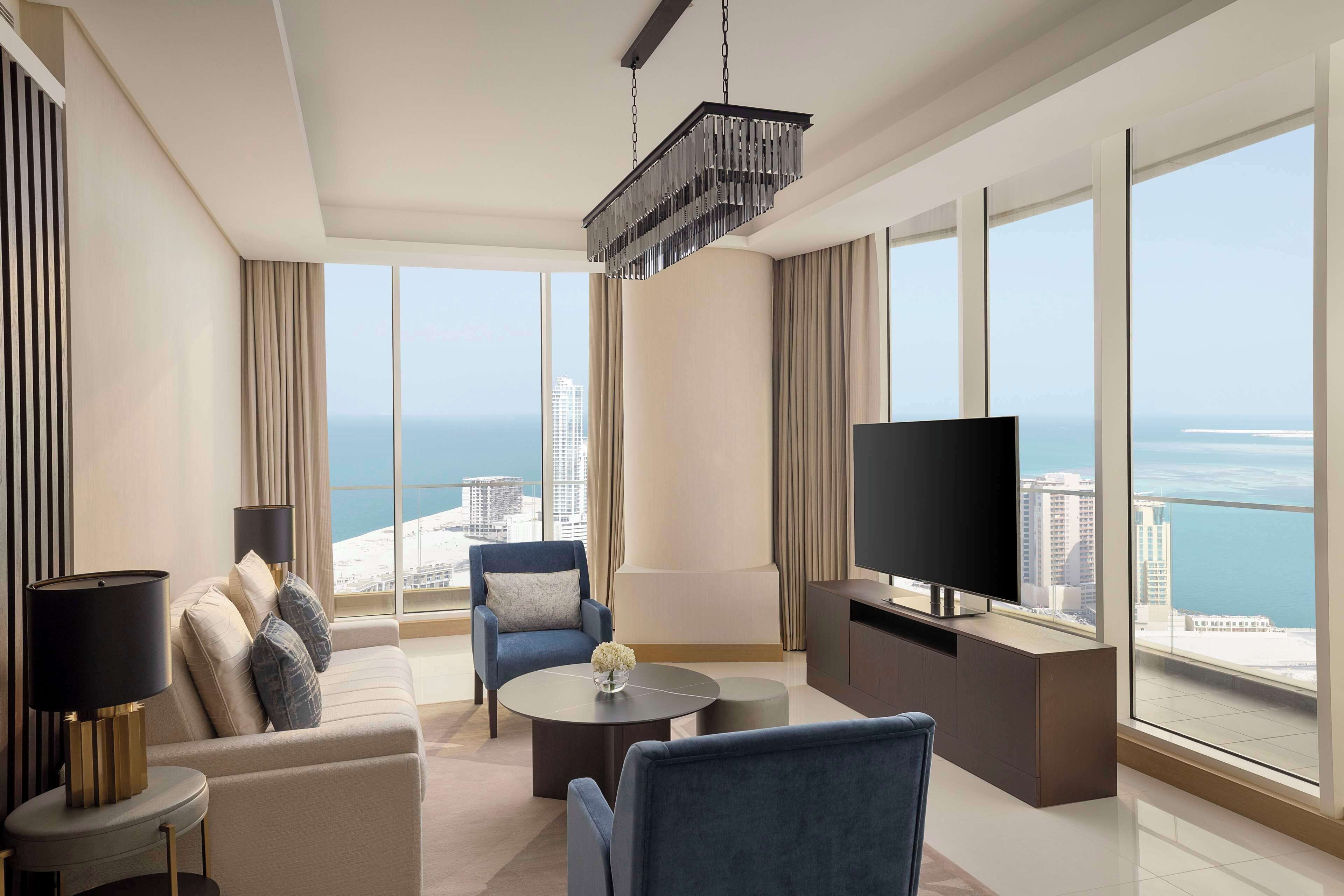 Un salon moderne comprenant un canapé, des fauteuils, une table basse et un téléviseur. De grandes fenêtres offrent une vue sur la ville et la mer.