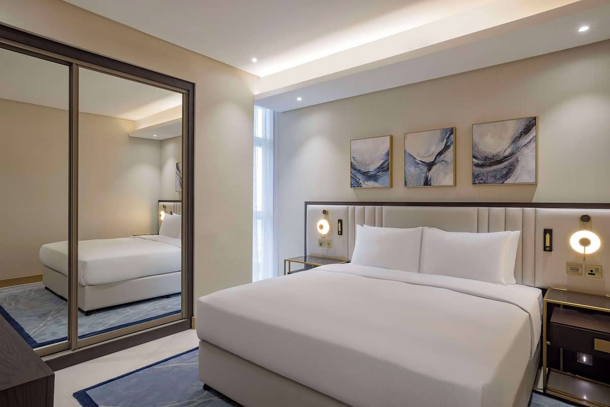 Habitación de hotel con una cama grande, una mesita de noche con lámpara, un espejo y arte abstracto en la pared.