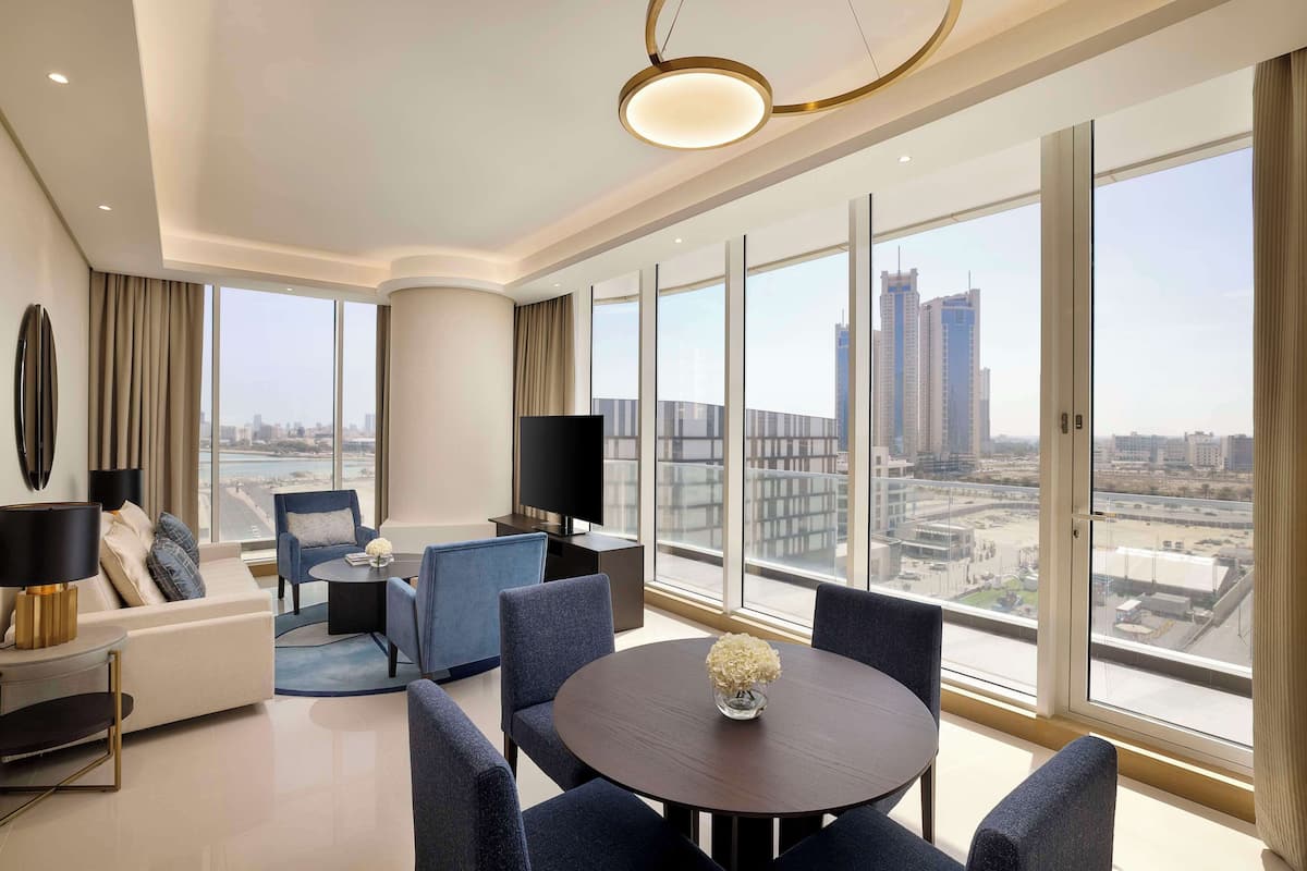 Una habitación de hotel moderna con un ventanal que ofrece vistas a la ciudad, un sofá, una mesa de comedor con sillas y un televisor de pantalla plana.