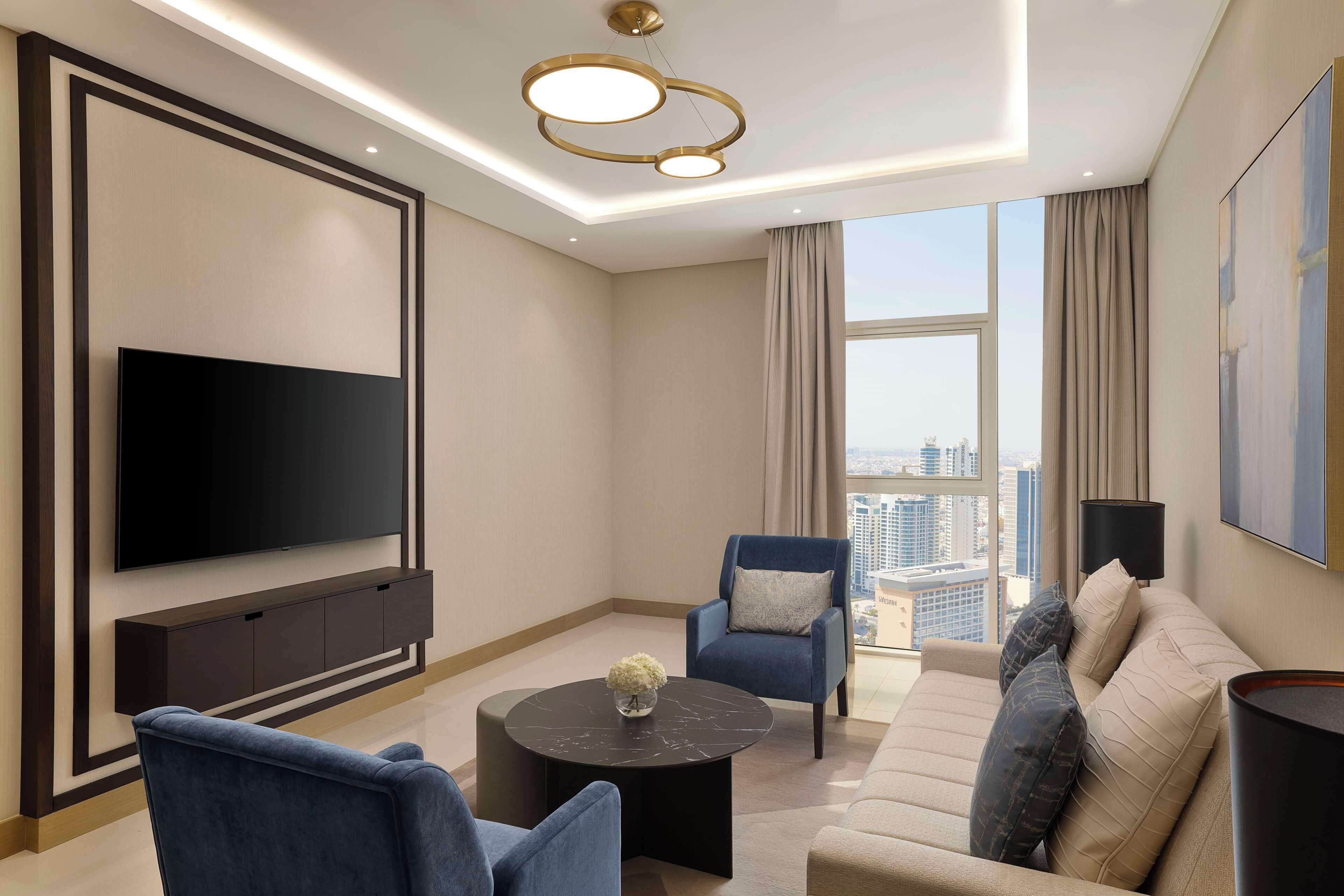 A modern living room with a flat-screen TV, a sofa, two armchairs, a coffee table, and a large window with a city view.