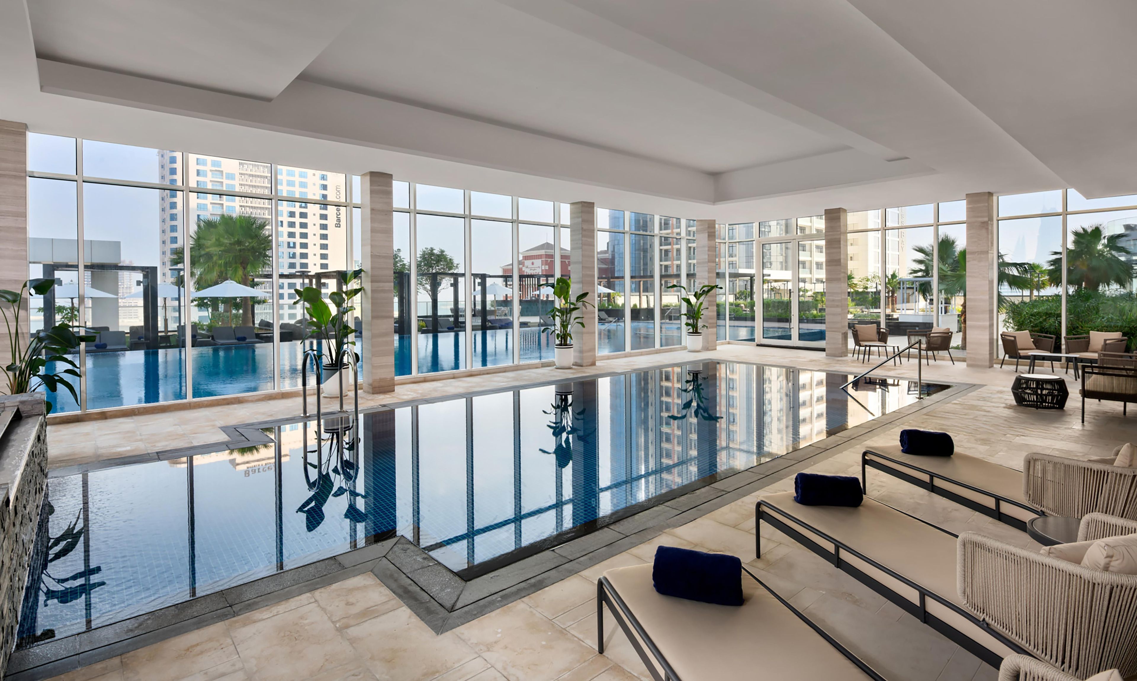 Una piscina interior moderna con paredes de vidrio transparente, zonas de descanso y plantas en macetas.