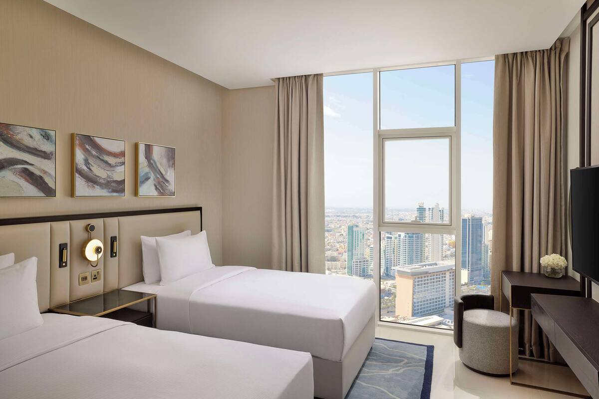 Habitación de hotel con dos camas, un escritorio, un televisor y un amplio ventanal con vista a la ciudad.