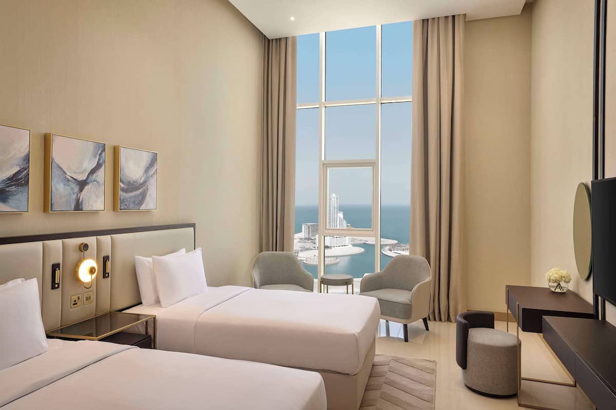Habitación de hotel con dos camas, un escritorio, una silla, un televisor y un ventanal grande con vistas a la ciudad y al agua.