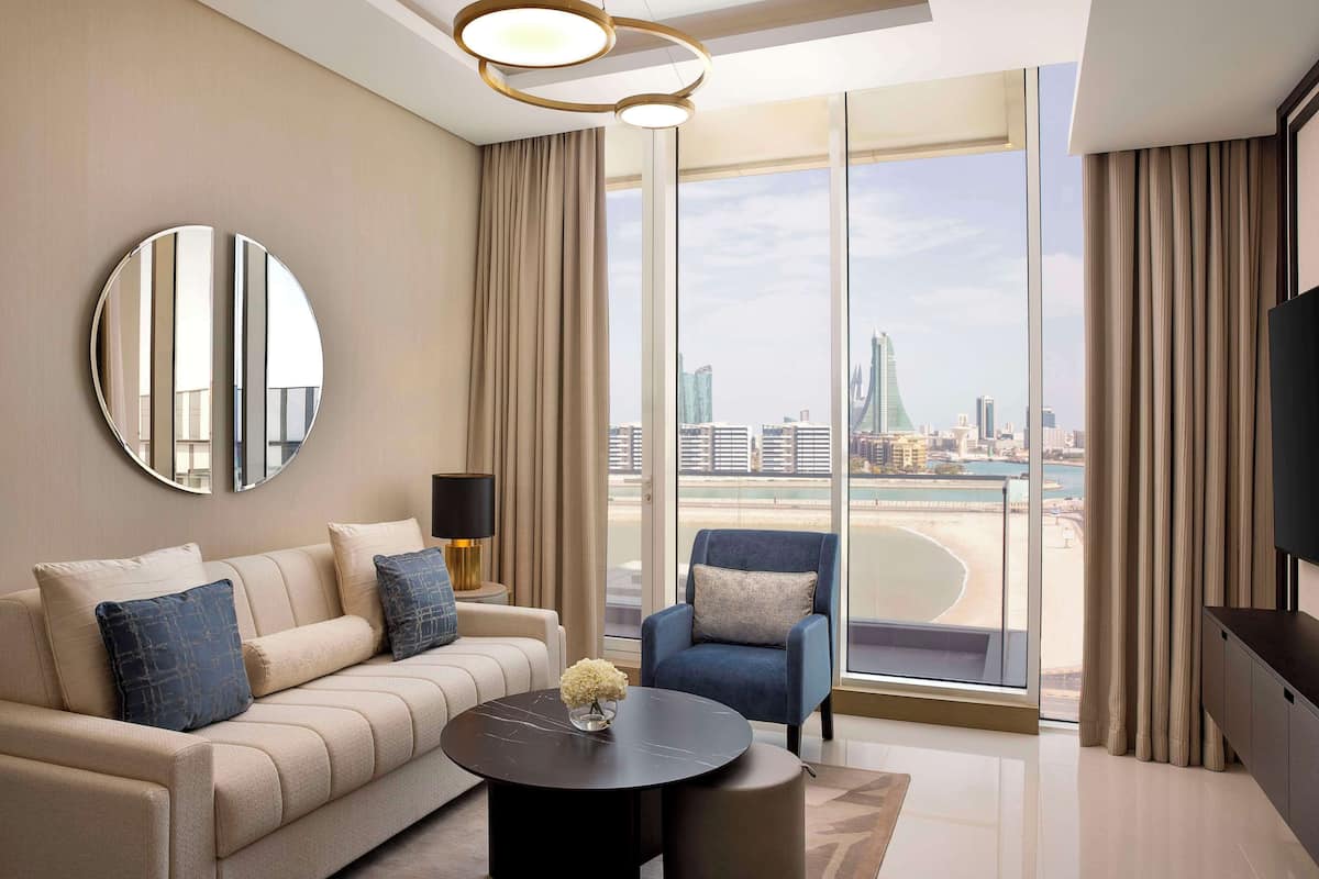 Un salón moderno con sofá, sillón y mesa de centro, ventanas amplias con vista a la ciudad y un espejo redondo en la pared.