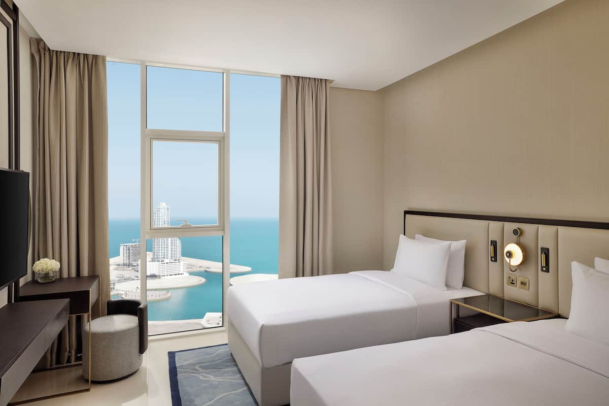 Habitación de hotel con dos camas, televisión, escritorio y vistas a la ciudad y al agua.