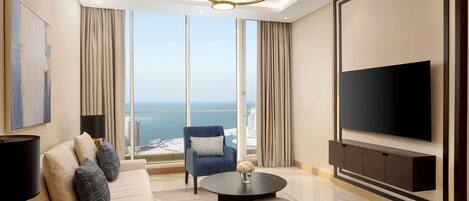 Un salón moderno con un televisor de pantalla plana grande, un sofá, una mesa de centro y vistas a la ciudad y al mar a través de amplios ventanales.