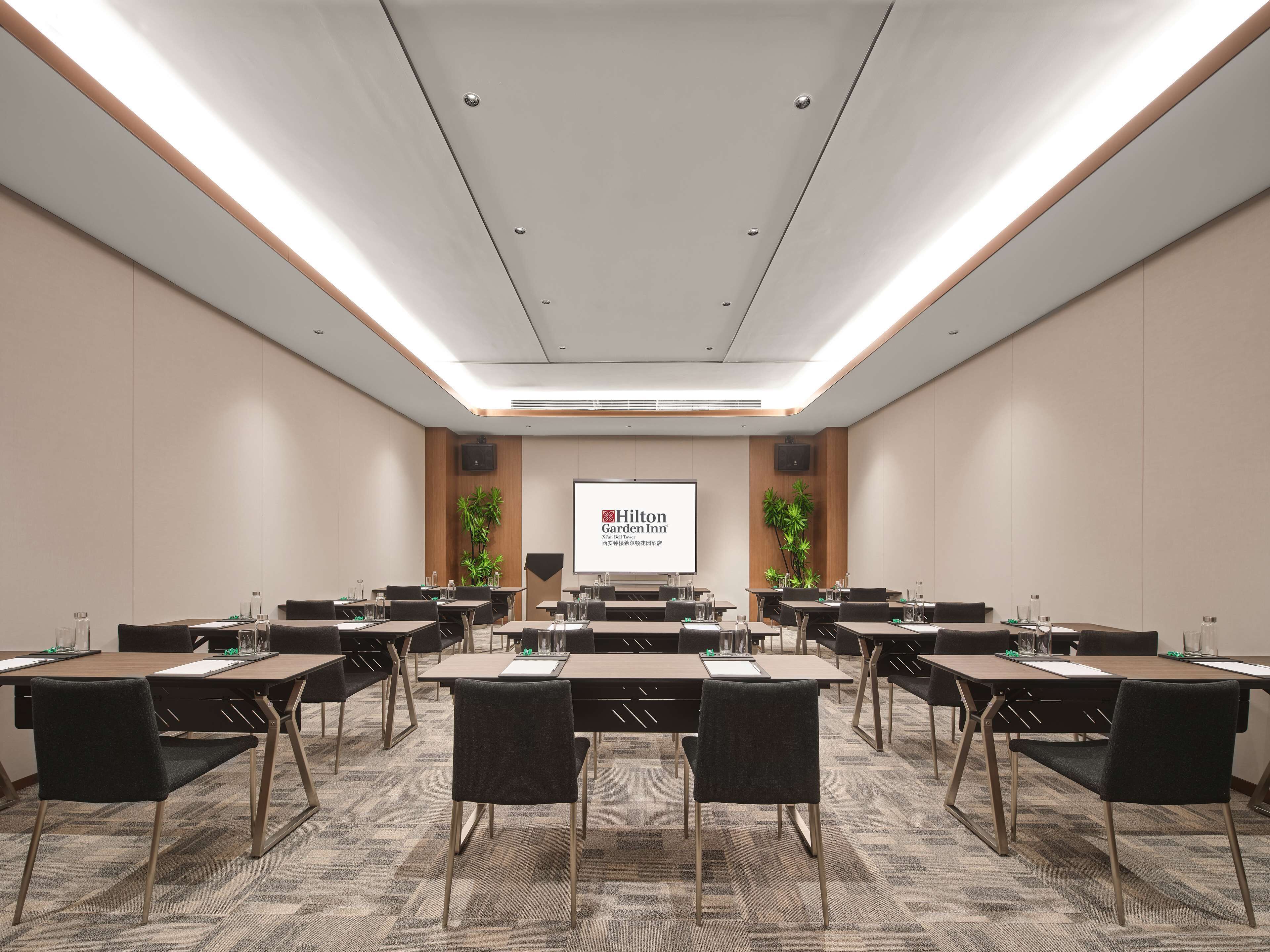 A conference room with multiple tables and chairs, a large screen displaying 'Hilton Garden Inn', and a wooden accent wall with plants.