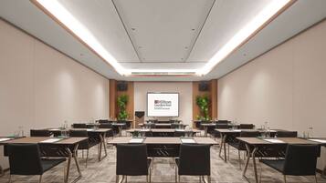 A conference room with multiple tables and chairs, a large screen displaying 'Hilton Garden Inn', and a wooden accent wall with plants.