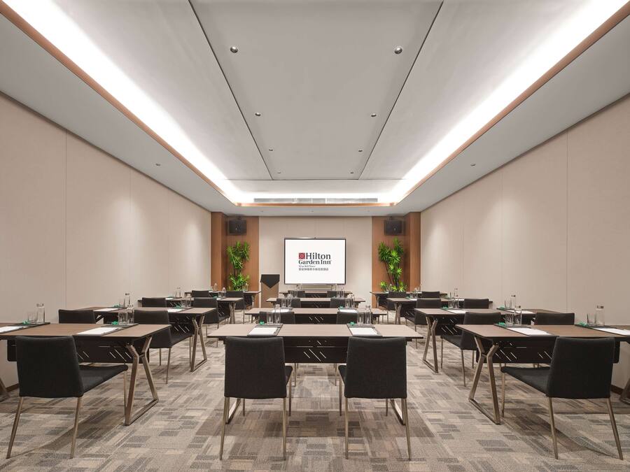 A conference room with multiple tables and chairs, a large screen displaying 'Hilton Garden Inn', and a wooden accent wall with plants.