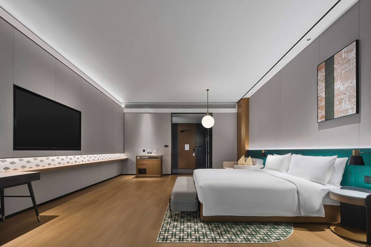 A modern hotel room with a large flat-screen TV, a bed with white bedding, a small desk with a chair, a bedside table, and a green accent wall.