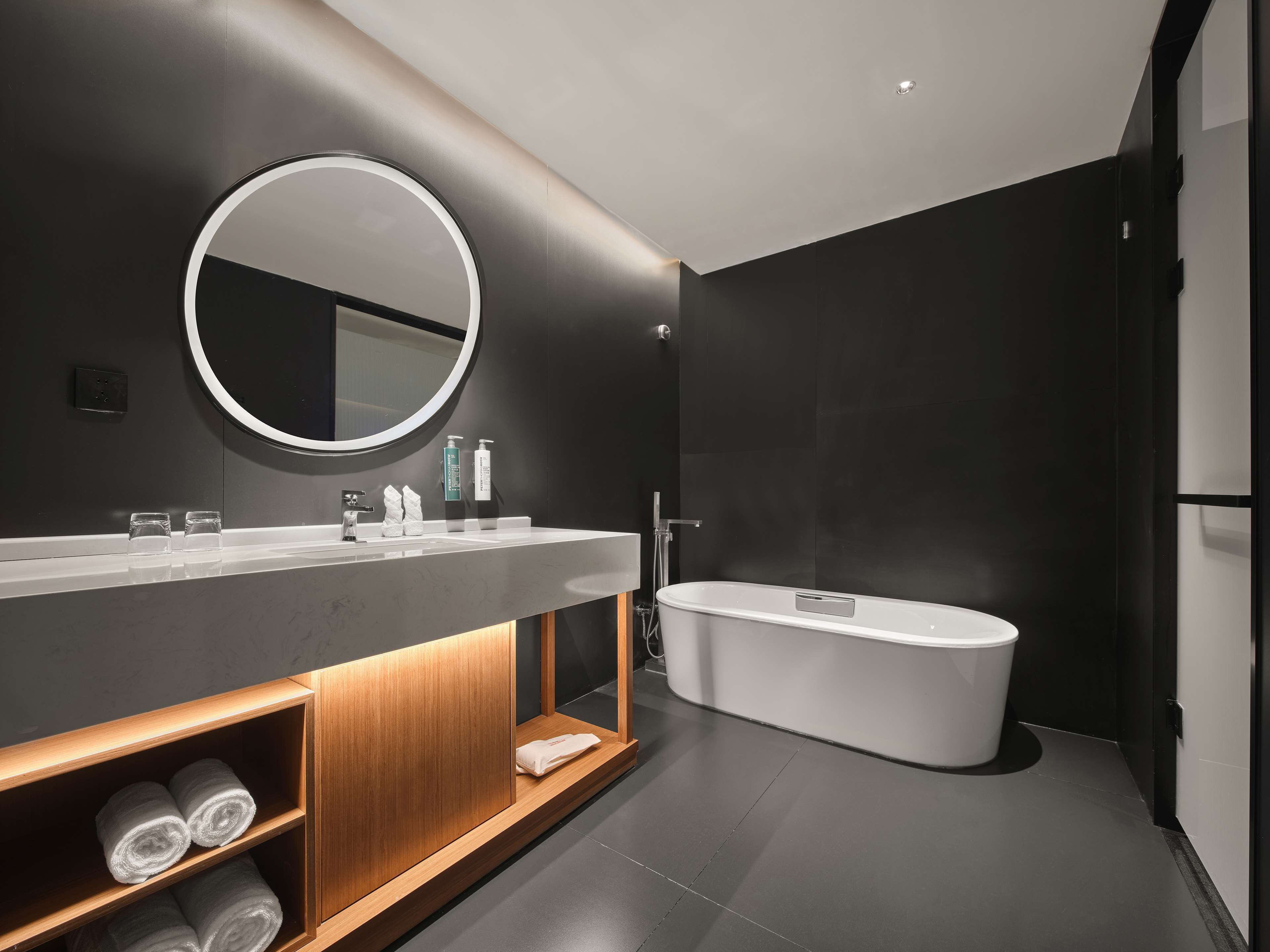 Un baño moderno con un espejo redondo, una bañera independiente y un mueble de tocador de madera con toallas.
