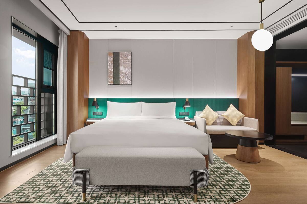 A modern hotel room with a large bed, a sofa, a small round table, and a view of the city through the window.