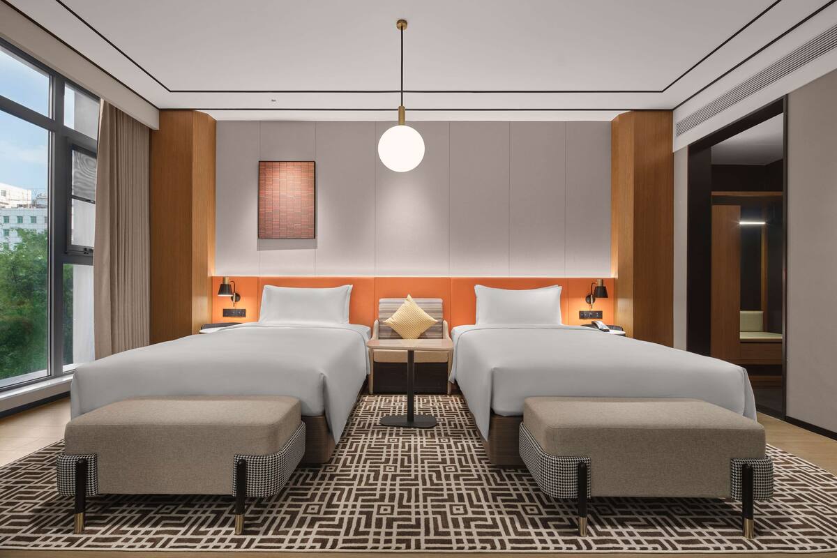 A modern hotel room with two beds, a large window, a patterned rug, and a wall-mounted light fixture.