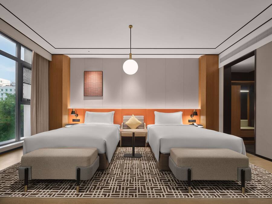 Een moderne hotelkamer met twee bedden, een groot raam, een kamerbreed tapijt met dessin en een wandlamp.