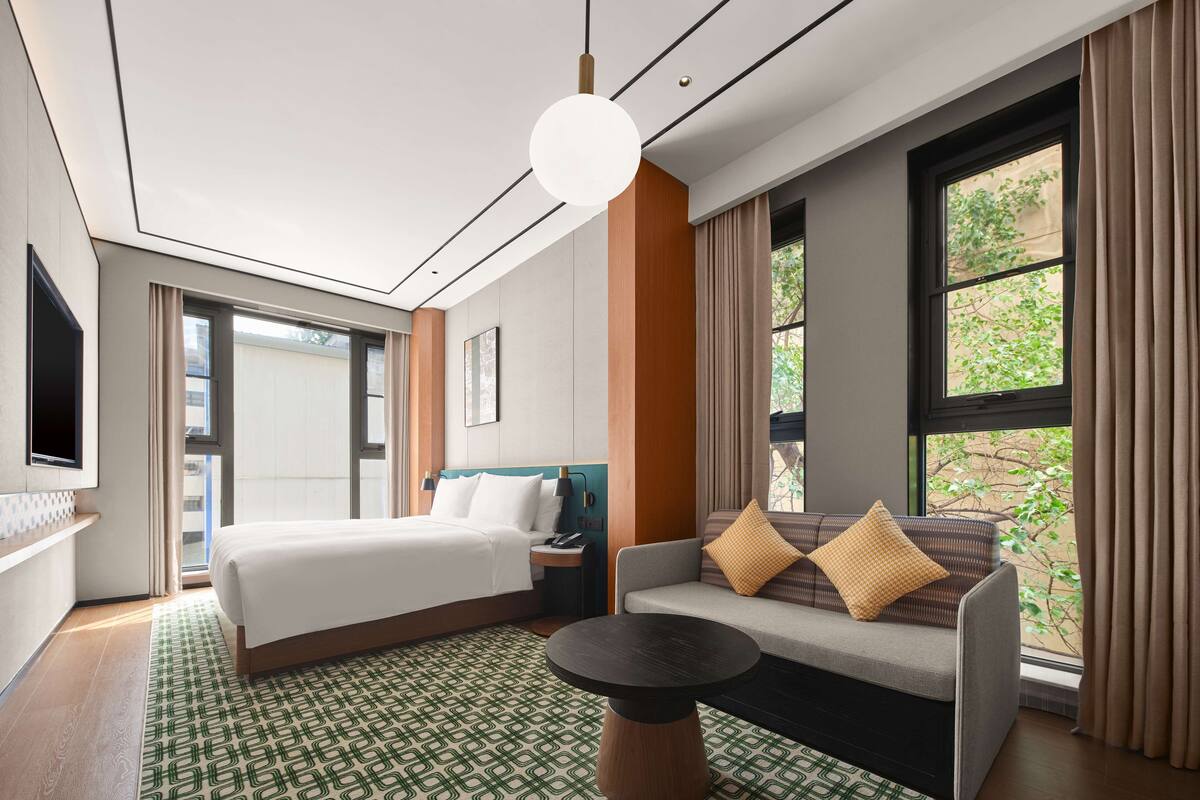 A modern hotel room with a large bed, a sofa with patterned cushions, a round coffee table, and a TV mounted on the wall.