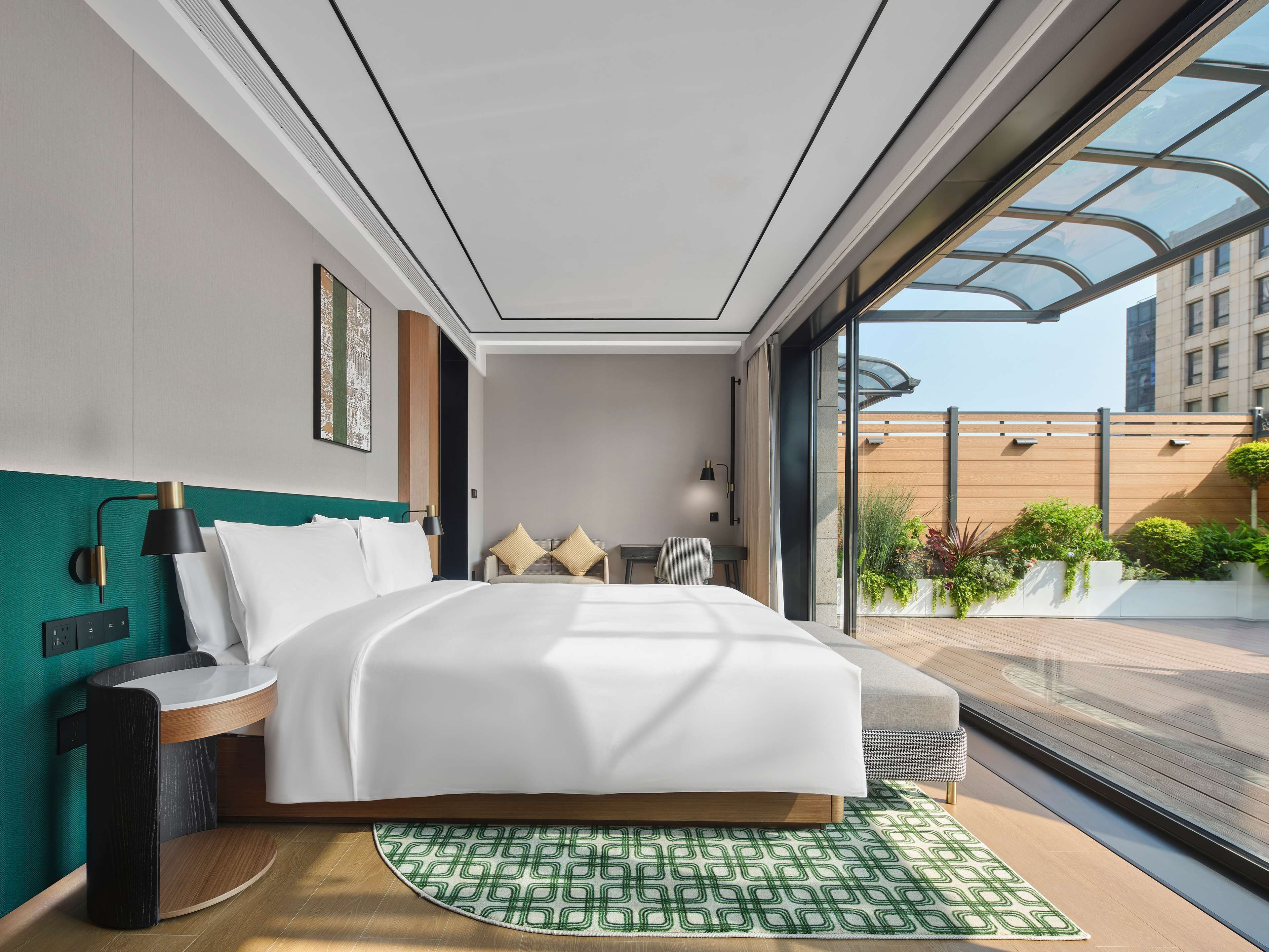 Une chambre d’hôtel moderne, dotée d’un grand lit, d’une table de chevet, d’une lampe et offrant une vue sur la ville grâce à de grandes fenêtres.