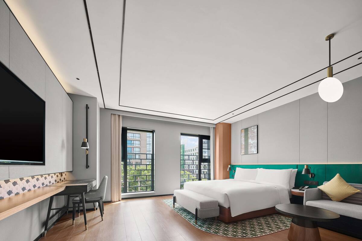 A modern hotel room with a large bed, a desk, a chair, and a sofa. There is a large window providing a view of buildings outside.