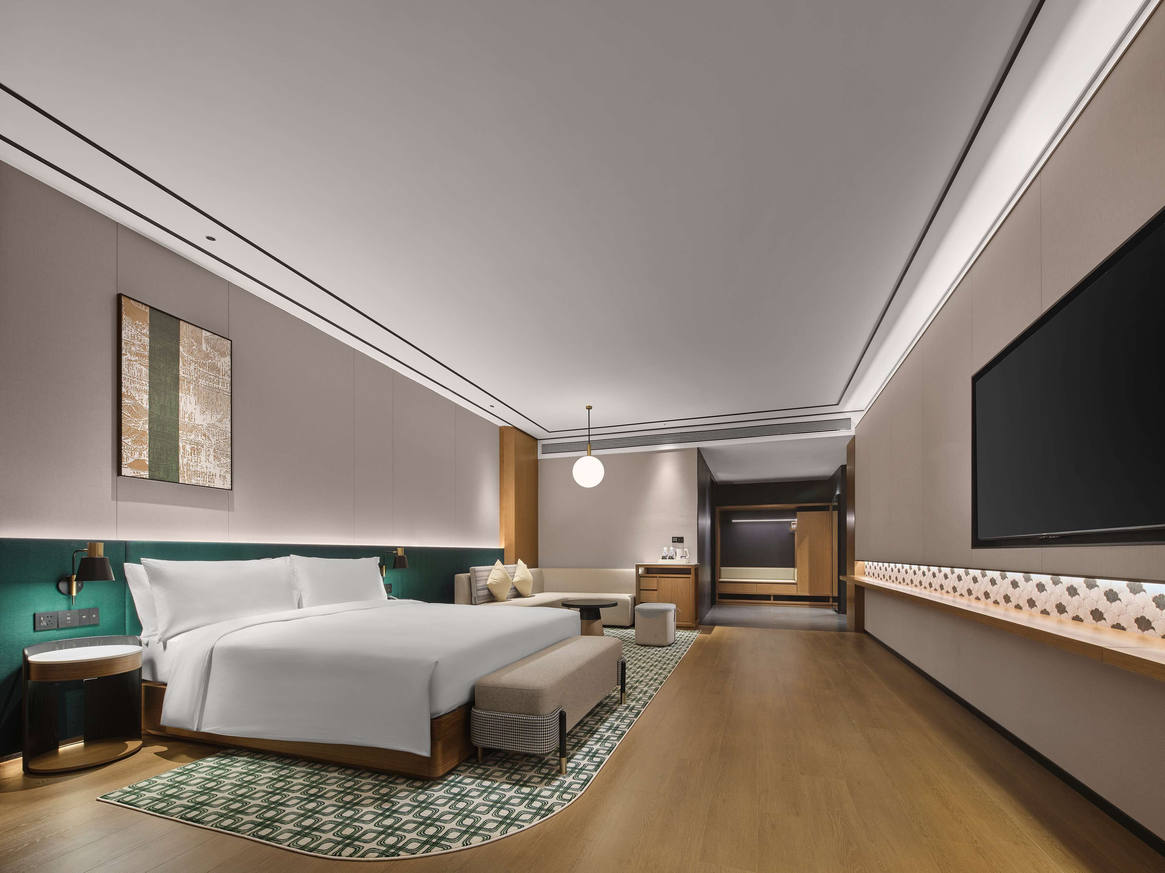 A modern hotel room with a bed, bedside table, lamp, and a large TV on the wall.
