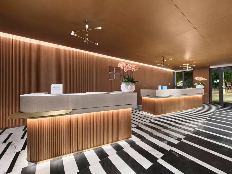 A modern hotel reception area with a wooden panel wall, a black and white checkered floor, and a central reception desk with a vase of pink flowers.