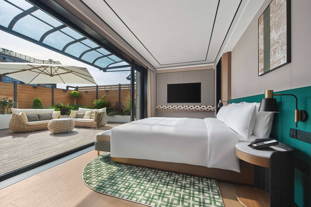 A modern hotel room with a large bed, a TV mounted on the wall, a bedside table with a lamp, and a view of the outdoor patio area.