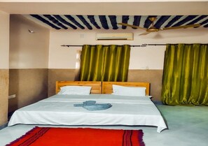 Un lit simple avec une tête de lit en bois, du linge de lit blanc et un tapis rouge. On peut voir un ventilateur de plafond et un appareil de climatisation.