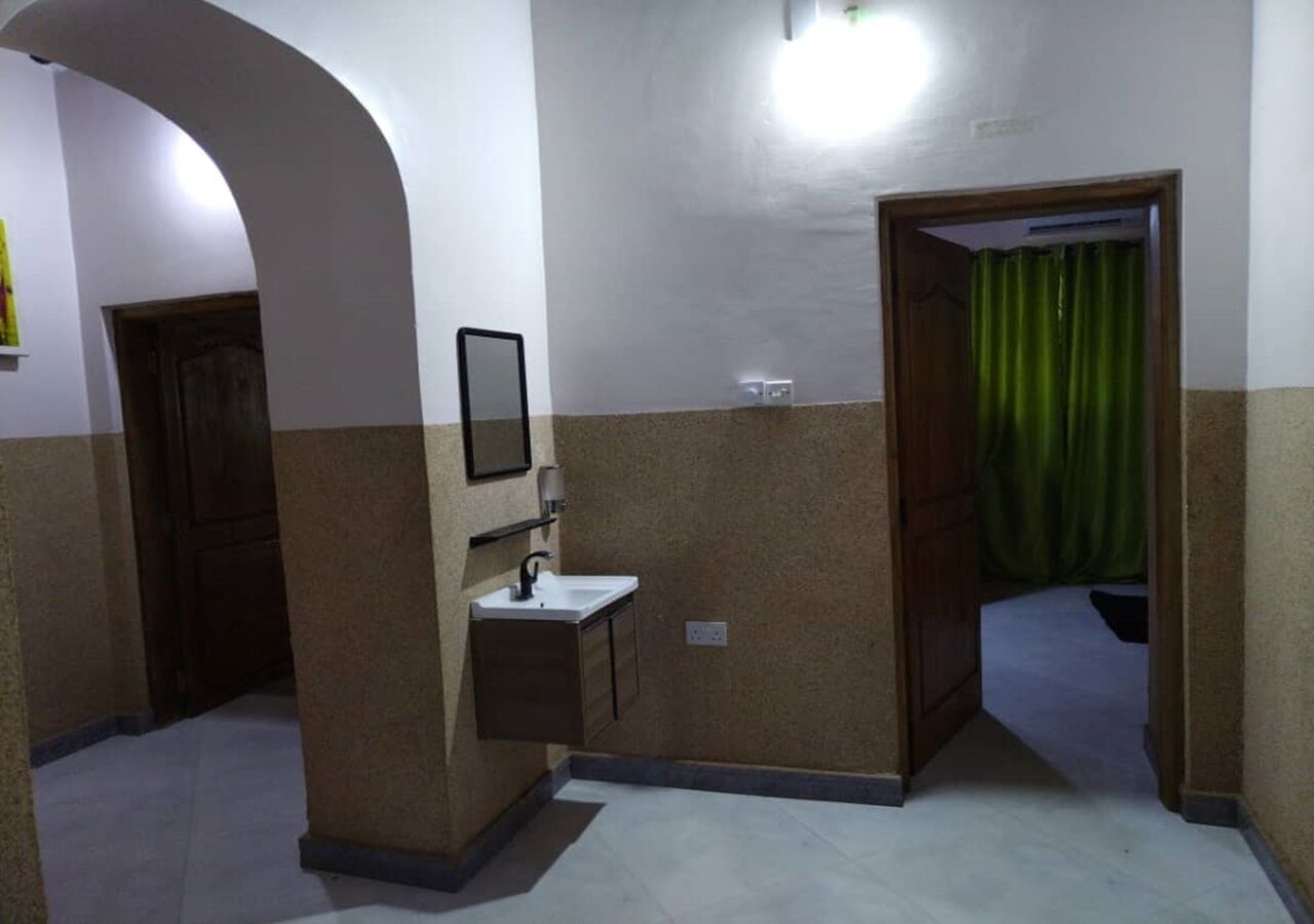A hallway with a sink and mirror, a door with a green curtain, and a light fixture on the wall.