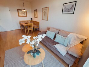 A living room with a brown sofa, a wooden dining table, and chairs. There is a vase with white flowers on a round table. - Vacation apartment "Ammersee" at Andechs Monastery (Andechs)