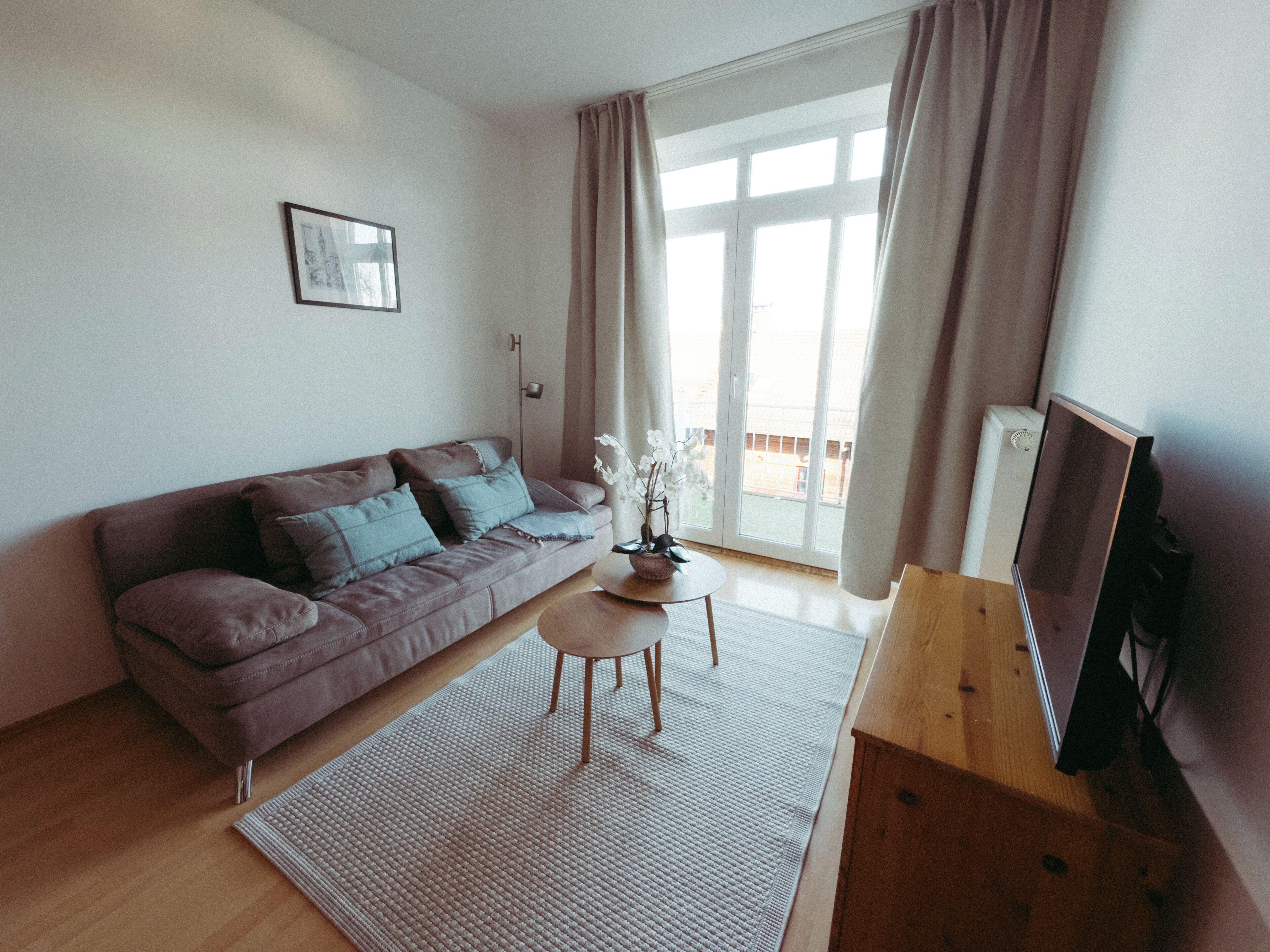Ein Wohnzimmer mit einer Couch, einem kleinen Tisch, einem Fernseher und einem Fenster mit Vorhängen.