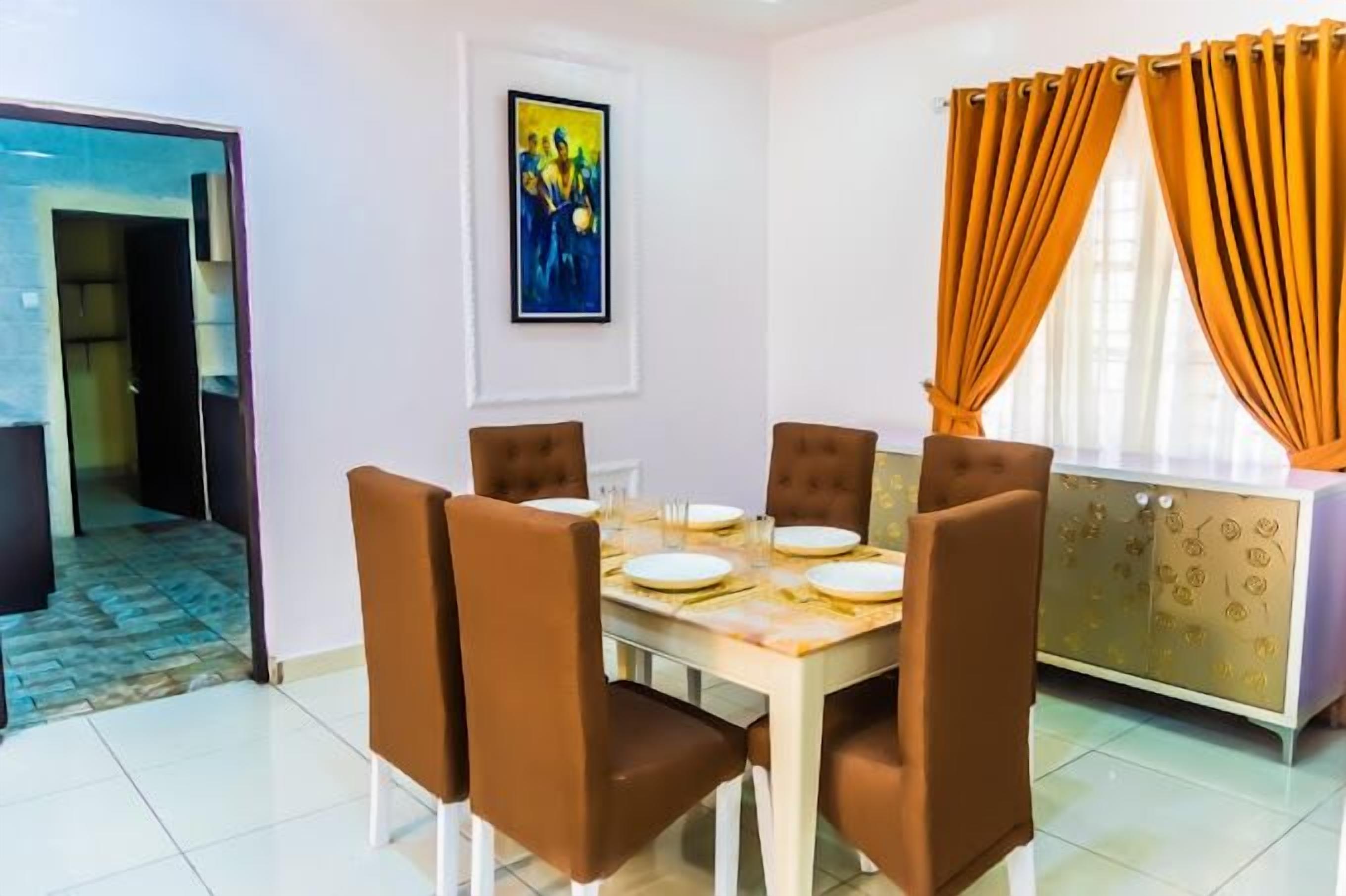Un comedor con una mesa dispuesta para cuatro, sillas color café tapizadas, un armario blanco con cubierta estampada y un cuadro en la pared.