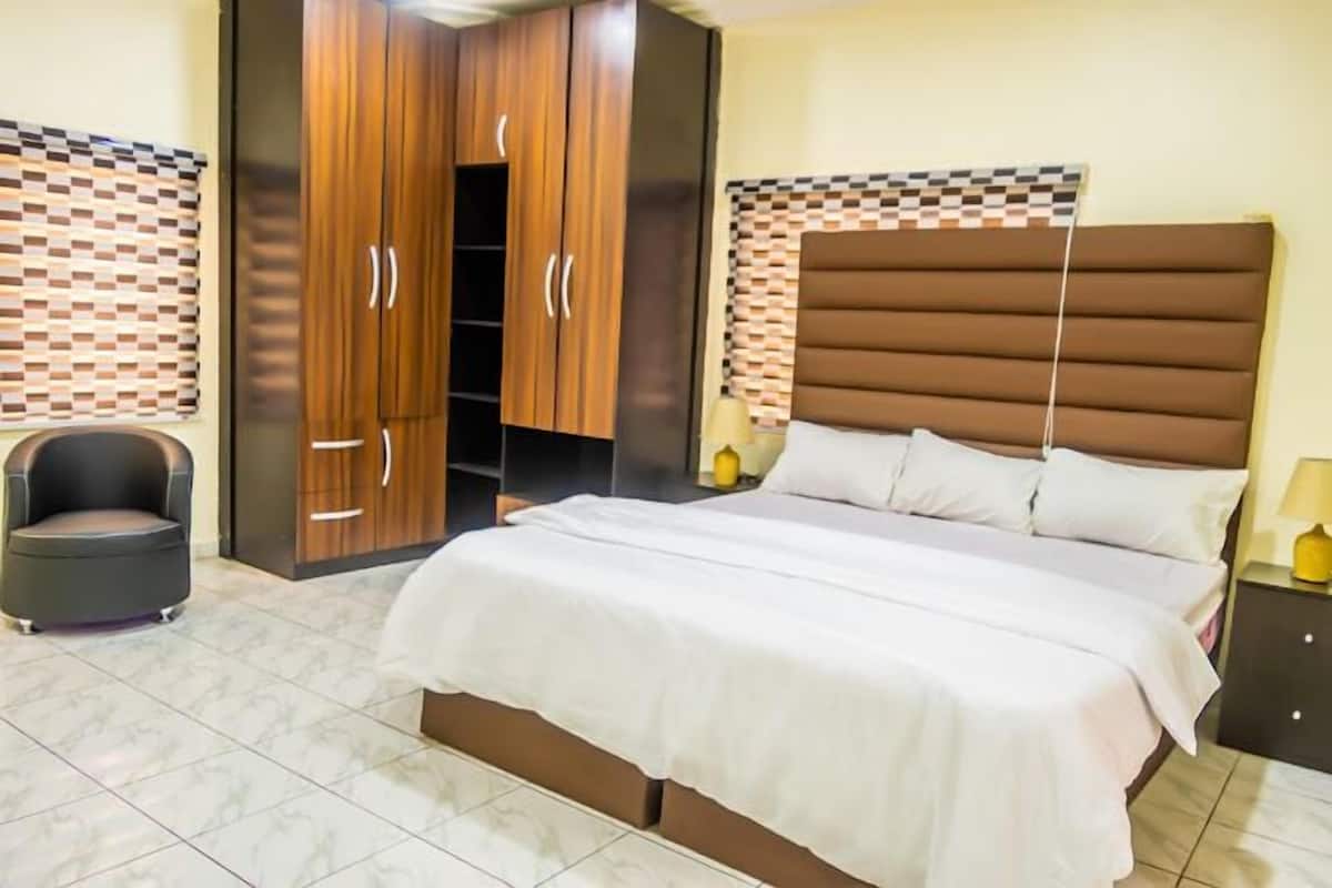 A bedroom with a large bed, wooden wardrobe, and a small chair.