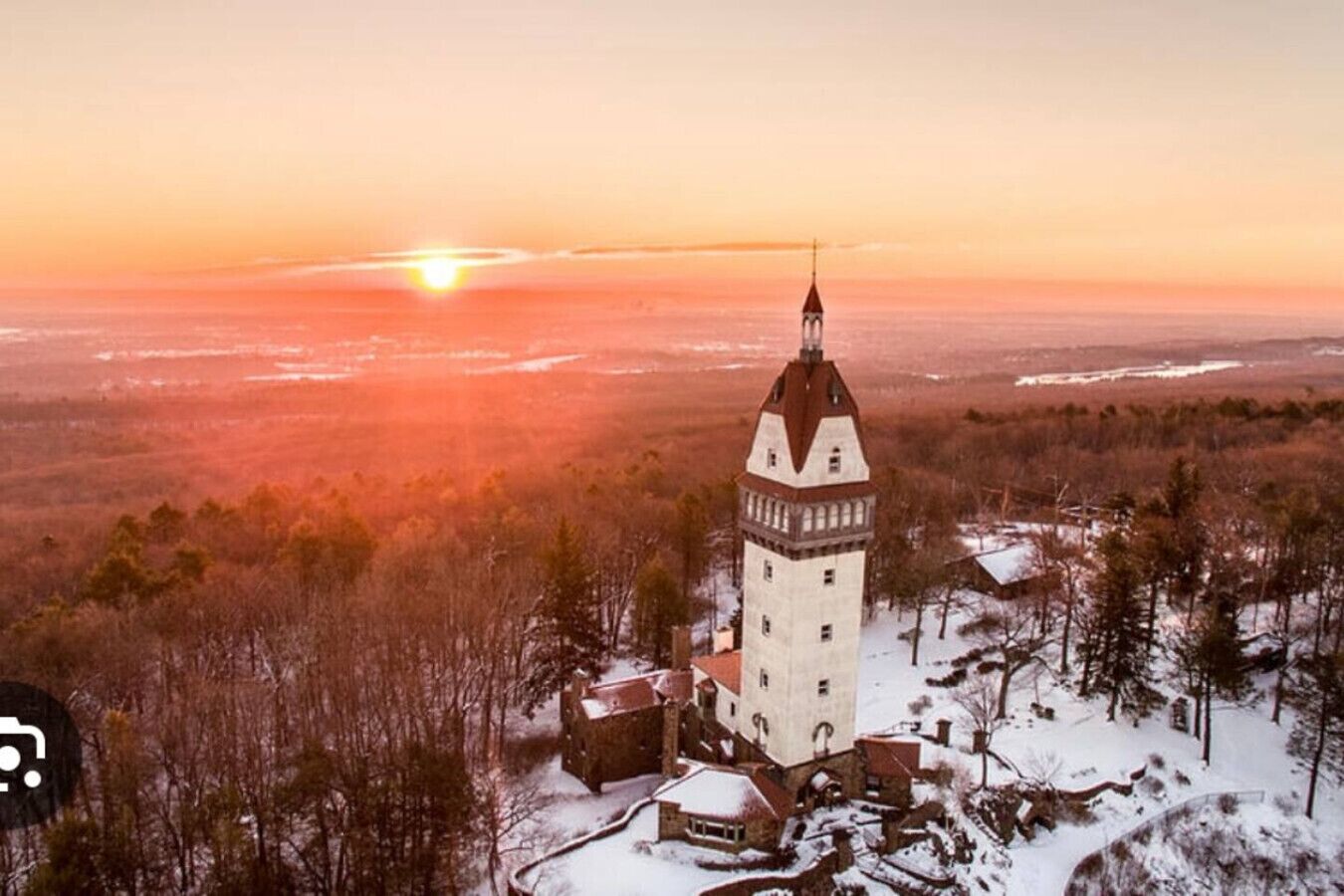 Una torre con aguja, rodeada de edificios y árboles cubiertos de nieve, durante el atardecer.