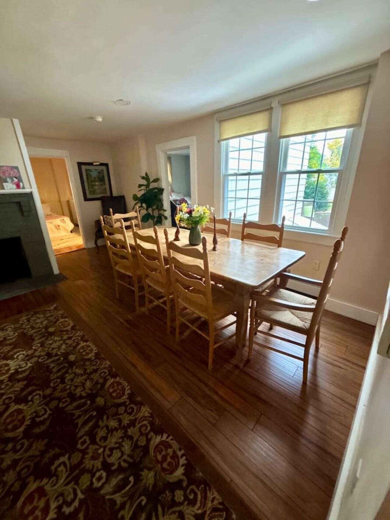 A dining area with a wooden table and chairs, a patterned rug, and a window with a view of greenery.