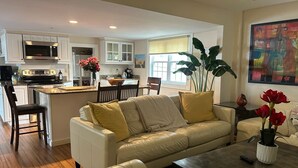 A modern kitchen with white cabinetry and a built-in microwave. There is a dining area with a white sofa and a coffee table. A large plant is in the corner, and a painting is on the wall. - The Old Avon Village (Avon)