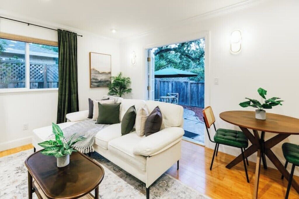A living room with a white sofa, a coffee table, and a dining table with chairs. There is a window with curtains, a plant on the coffee table, and a view of the backyard through an open door.