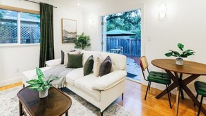 A living room with a white sofa, a coffee table, and a dining table with chairs. There is a window with curtains, a plant on the coffee table, and a view of the backyard through an open door. - Serene Escape 2BD /w Private Patio near Stanford (Menlo Park)