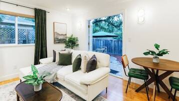 A living room with a white sofa, a coffee table, and a dining table with chairs. There is a window with curtains, a plant on the coffee table, and a view of the backyard through an open door.