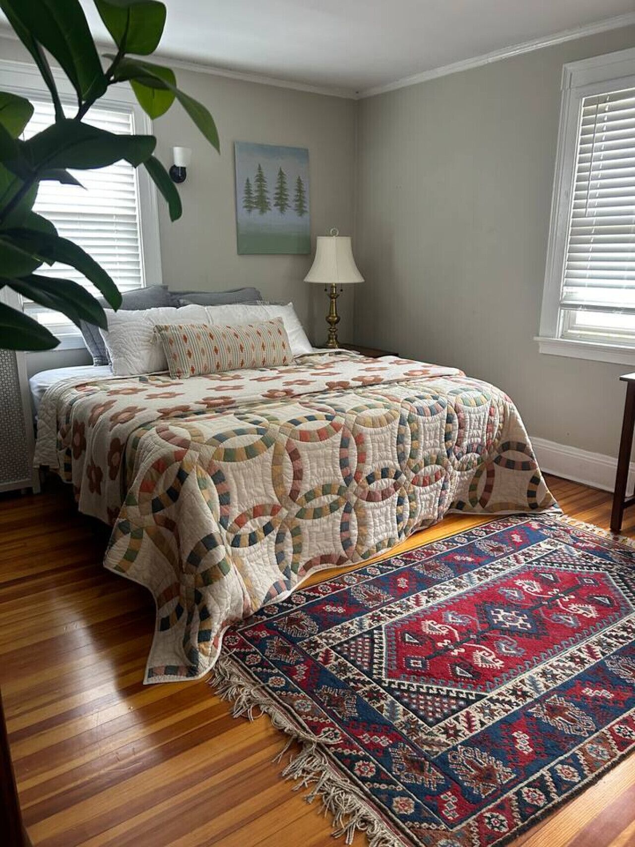 A bedroom with a bed, a lamp, a painting, and a large plant.
