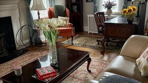 Een gezellige woonkamer met een open haard, een bijzettafeltje met een boek, een bank en een eettafel met zonnebloemen.