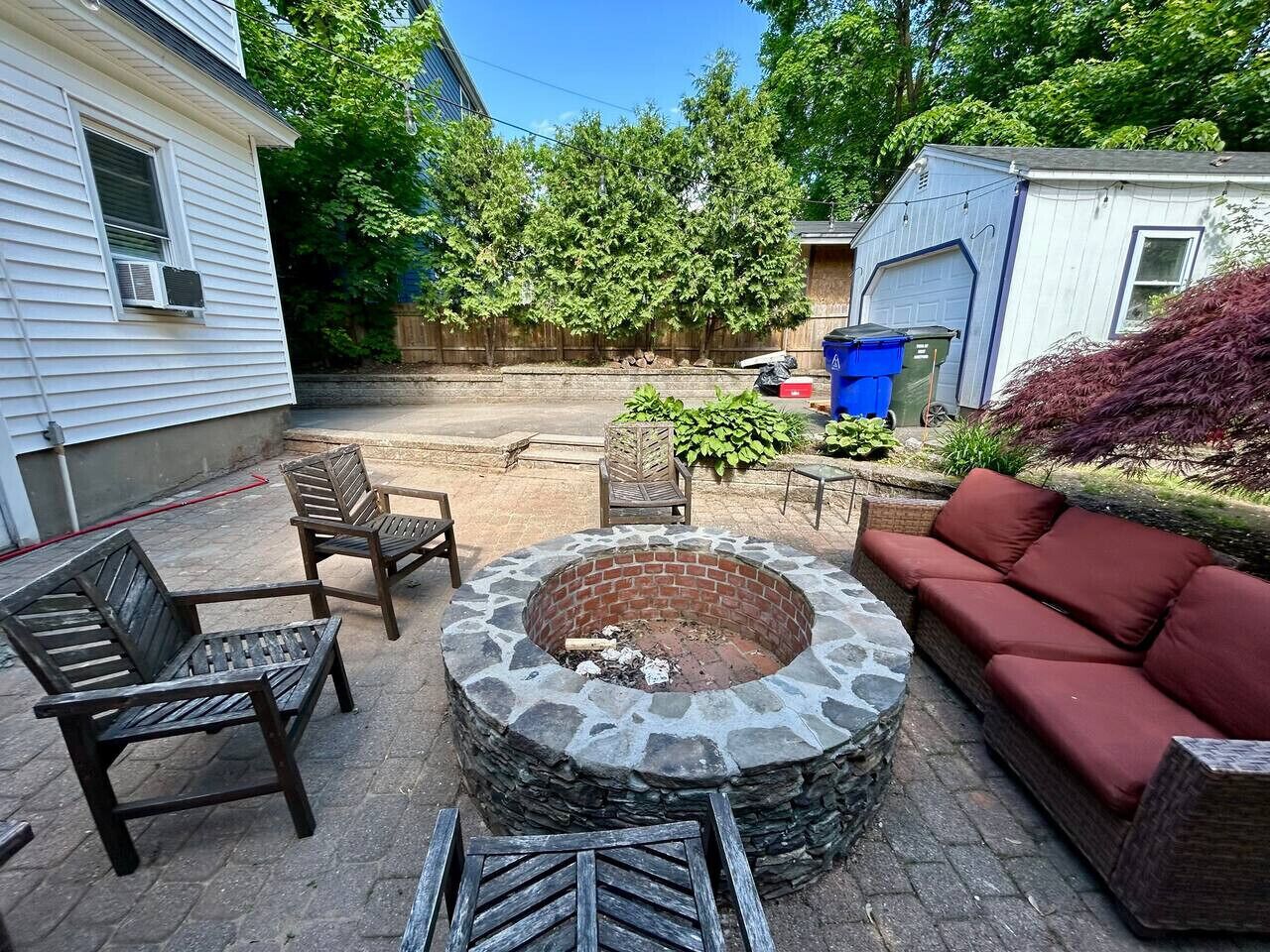 A backyard with a stone fire pit, wooden chairs, a red sofa, and a shed in the background.