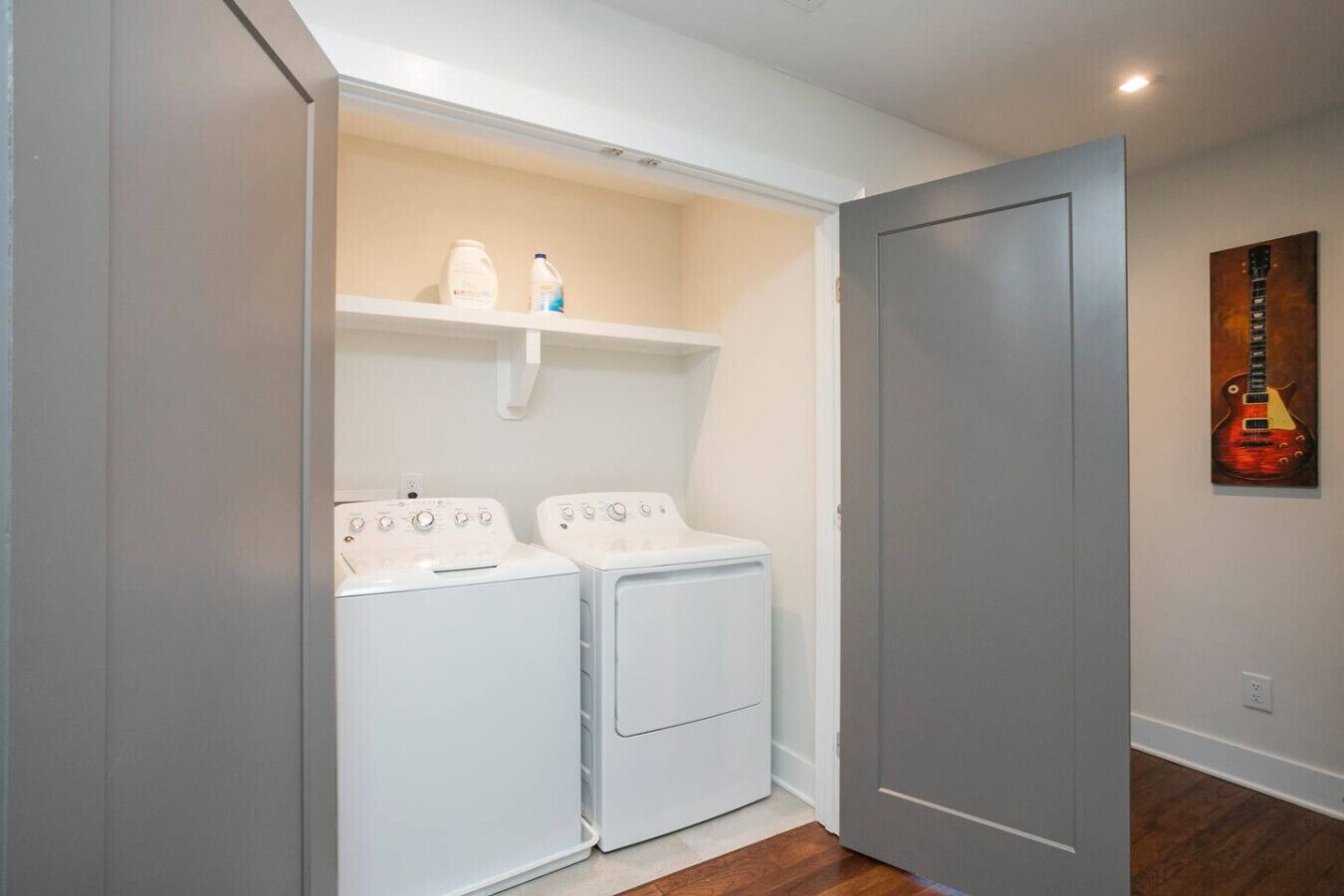 Una sala de lavandería con dos lavadoras y secadoras de carga frontal blancas, un estante superior con botellas de detergente, y una puerta a la derecha.