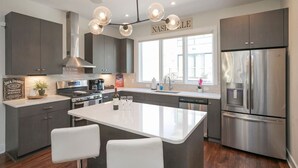 A modern kitchen with grey cabinets, a stainless steel refrigerator, a large island with bar stools, and a hanging light fixture. - Luxury Escape | Spacious & Safe, 7min to Downtown! (Nashville)