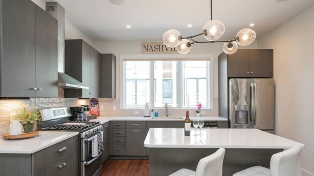 A modern kitchen with dark cabinets, a central island with white bar stools, stainless steel appliances, and a pendant light fixture. - Luxury Escape | Spacious & Safe, 7min to Downtown! (Nashville)